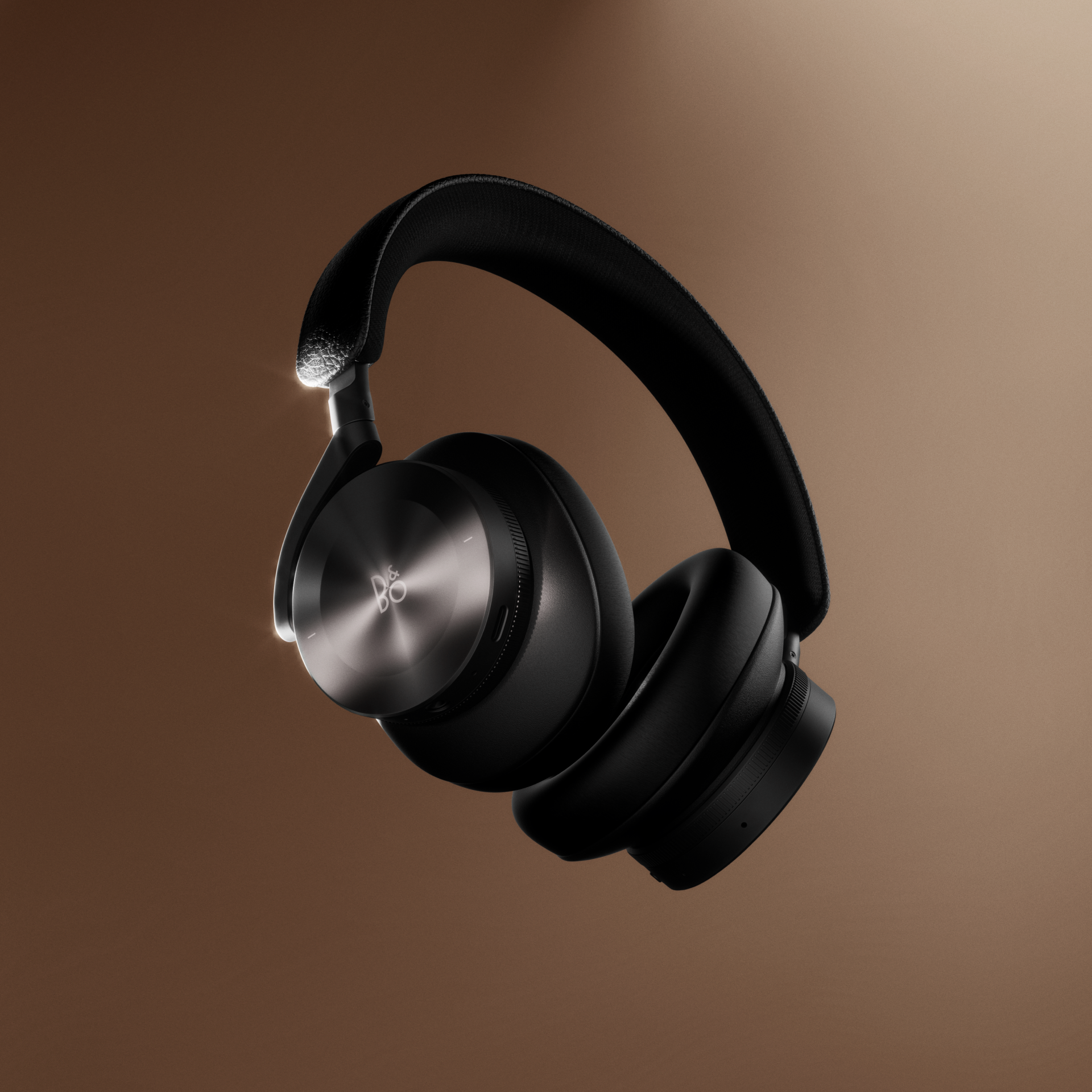 Schwebende Studio-Aufnahme der Bang & Olufsen Beoplay H95 Over-Ear-Kopfhörer in Black Anthracite. Die Ansicht betont das ikonische Radial-Finish der Aluminium-Ohrmuscheln und das hochwertig texturierte Leder-Kopfband vor einem warmen, bronze-braunen Farbverlauf.