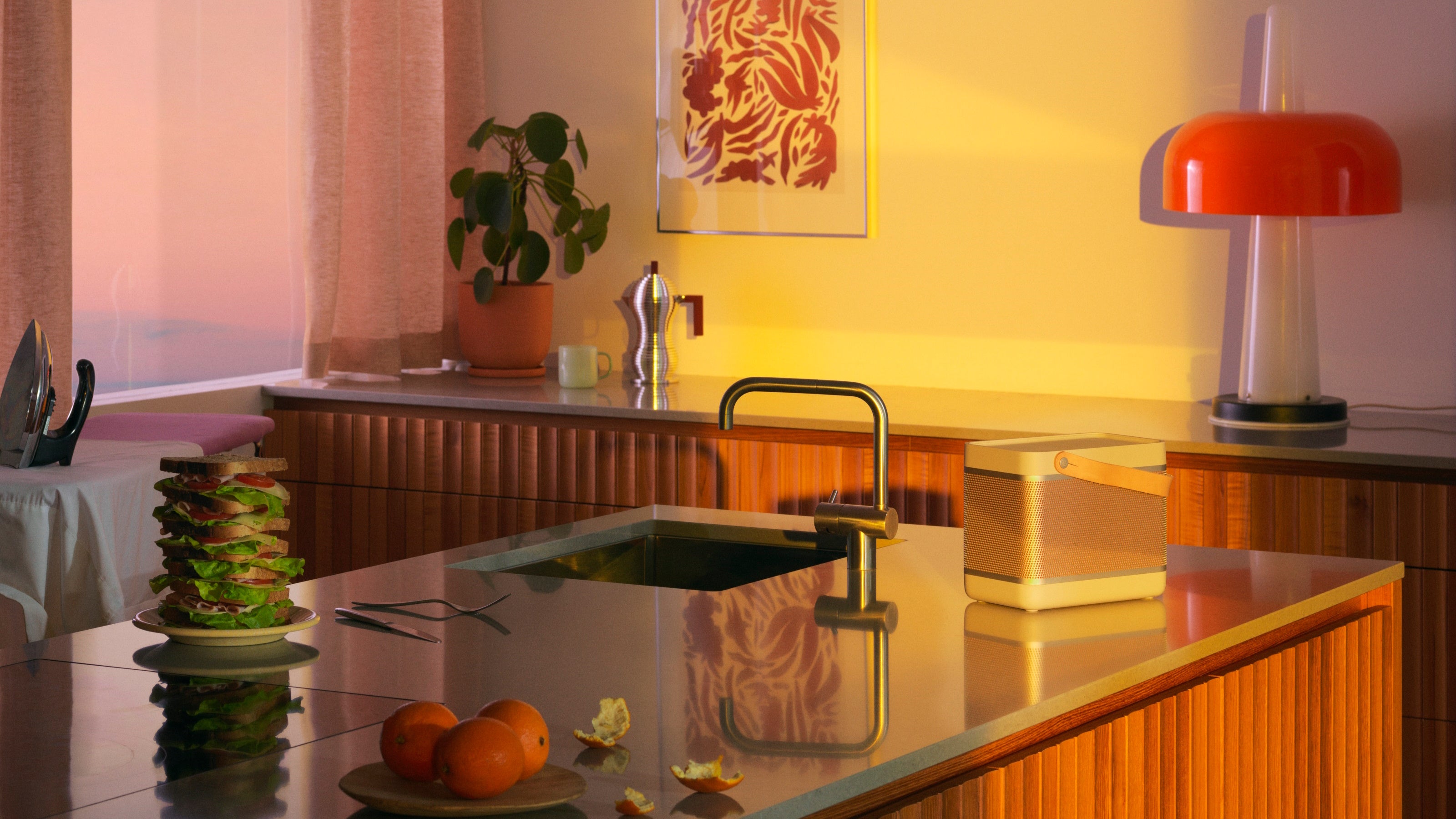 Stimmungsvolle Lifestyle-Aufnahme des Bang & Olufsen Beolit 20 Lautsprechers auf einer modernen Kücheninsel mit Holzfronten und Marmorarbeitsplatte. Die Szene wird von warmem, goldenem Licht durchflutet und zeigt eine rote Pilz-Tischlampe, eine Espressokanne und einen imposanten Sandwich-Turm im Hintergrund.