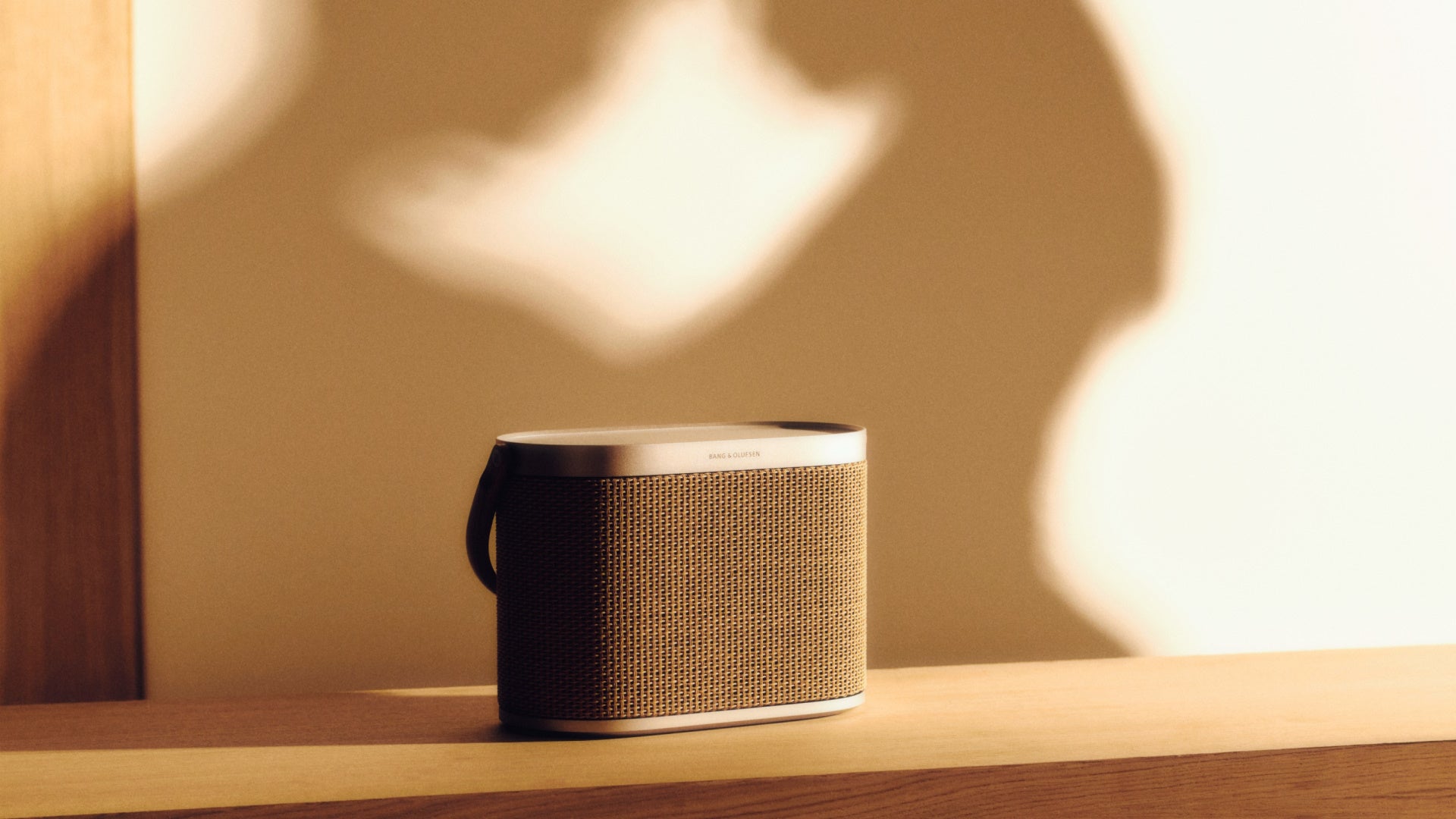 Stimmungsvolle Produktaufnahme des Bang & Olufsen Beosound A5 Heimlautsprechers auf einem Holzboard. Das geflochtene Cover aus Papierfasern und der Rahmen aus hellem Aluminium werden durch ein Spiel aus warmem Sonnenlicht und weichen Schatten an der Wand kunstvoll in Szene gesetzt.