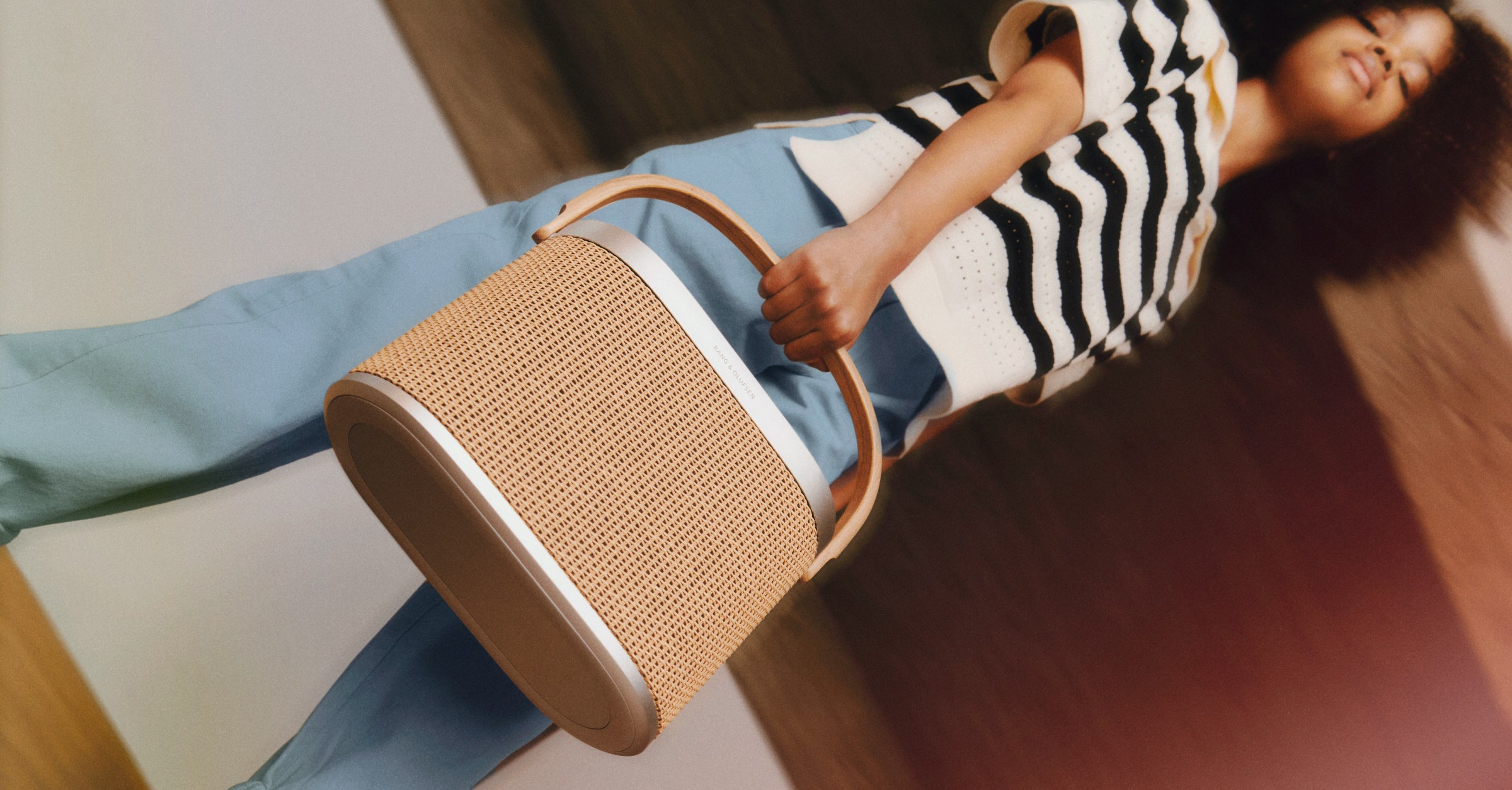 Modische Lifestyle-Aufnahme des portablen Bang & Olufsen Beosound A5 Lautsprechers. Eine Person trägt den Lautsprecher am markanten hellen Holzgriff. Das Design besticht durch ein geflochtenes Papiergeflecht-Cover und einen Rahmen aus silbernem Aluminium, was die Ästhetik eines hochwertigen Design-Accessoires unterstreicht.