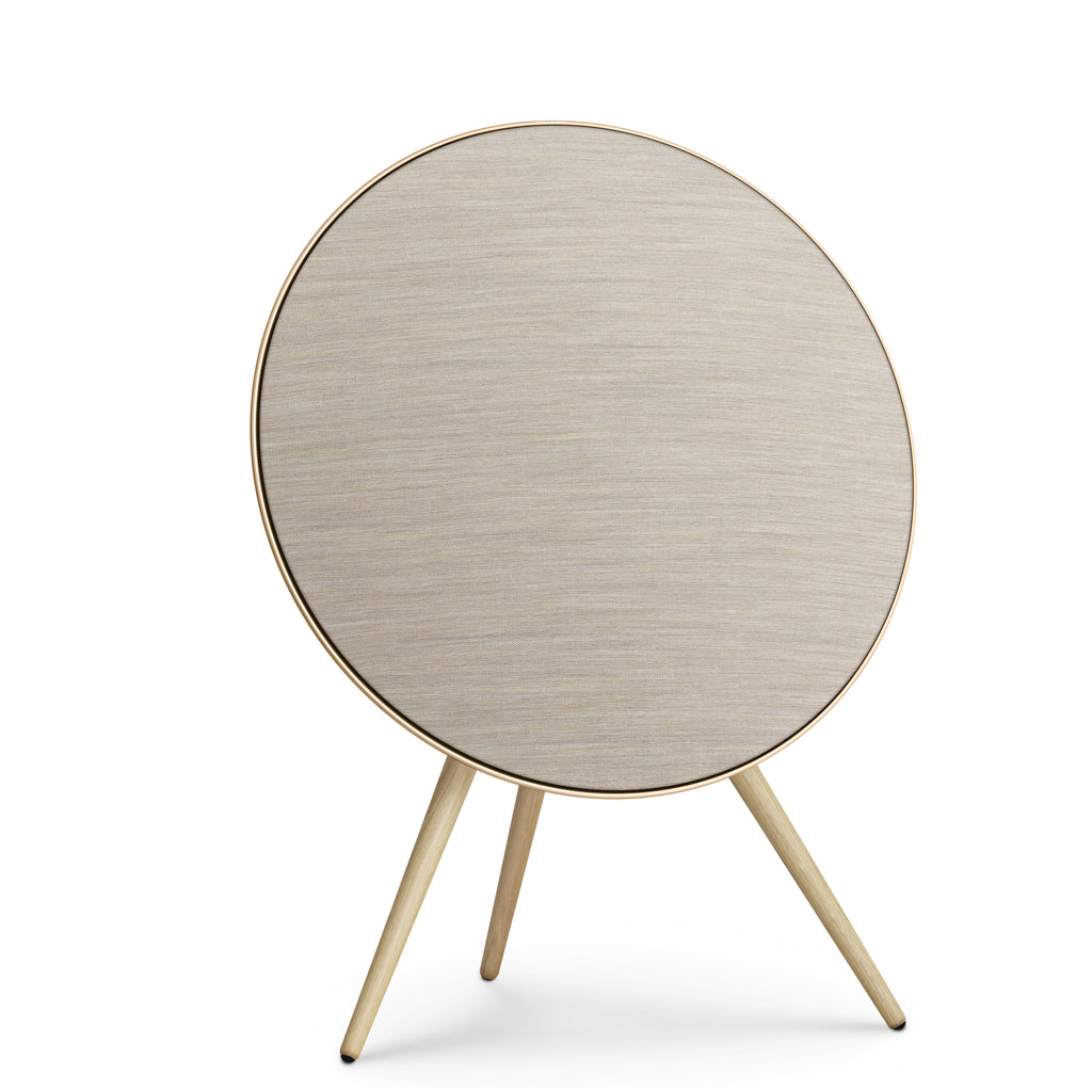 Frontansicht der Beosound A9 in Gold Tone. Das kreisrunde Design mit sandfarbenem Kvadrat-Bezug und hellen Eichenholzbeinen wirkt wie ein zeitloses Kunstobjekt.