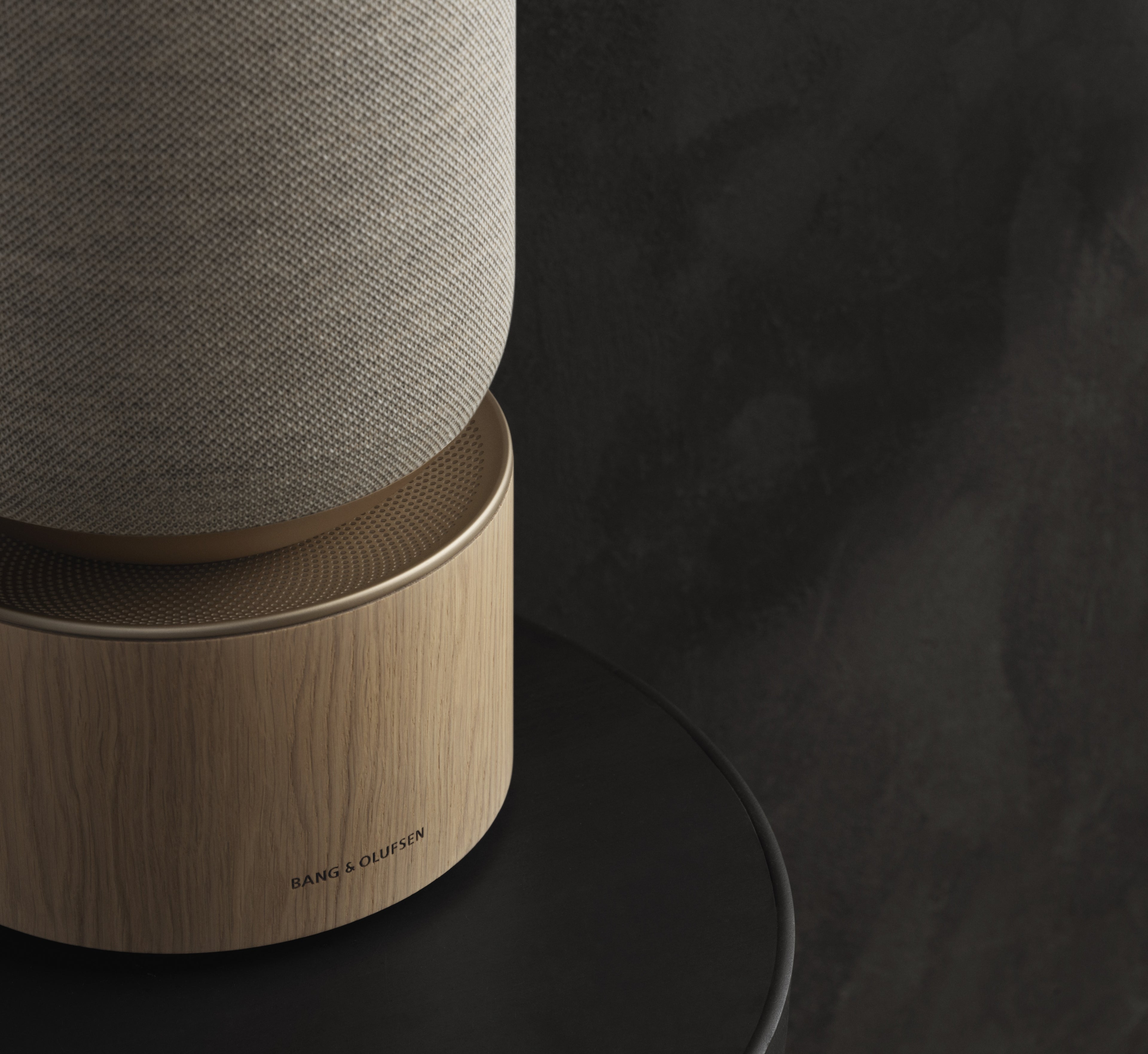 Nahaufnahme des Bang & Olufsen Beosound Balance Heimlautsprechers. Das Bild zeigt den Sockel aus hellem Eichenholz mit dezentem B&O-Logo, darüber die fein gelochte Aluminium-Trennschicht in Gold-Tone und den oberen Teil mit einer hochwertigen Stricktextil-Bespannung vor einem dunklen Hintergrund.