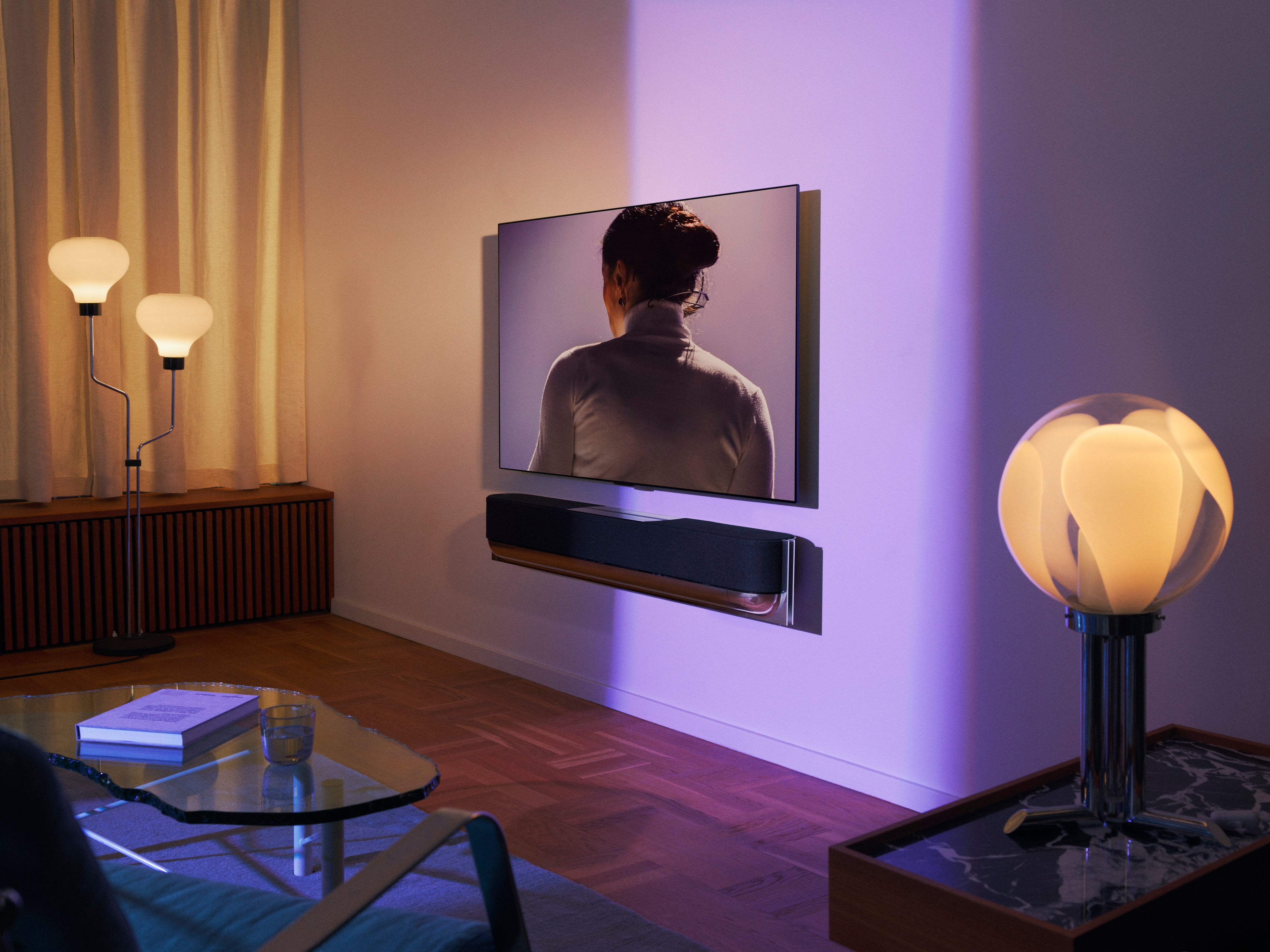 Bang & Olufsen Beovision Theatre in der Wandmontage-Konfiguration mit eleganter, dunkler Textilfront. Die Lifestyle-Aufnahme zeigt das schwebende Design des Heimkinosystems an einer weißen Wand, akzentuiert durch violettes Licht in einem luxuriös eingerichteten Wohnzimmer.