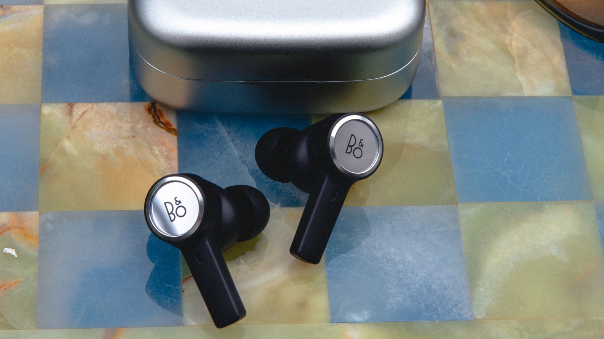 Draufsicht auf die Bang & Olufsen Beoplay Eleven In-Ear-Kopfhörer in Black Anthracite. Die Earbuds mit markantem silbernem Aluminium-Ring und Logo-Prägung liegen vor ihrem massiven, passenden Aluminium-Ladecase auf einer blau-gelben Marmor-Oberfläche im Schachbrettmuster.