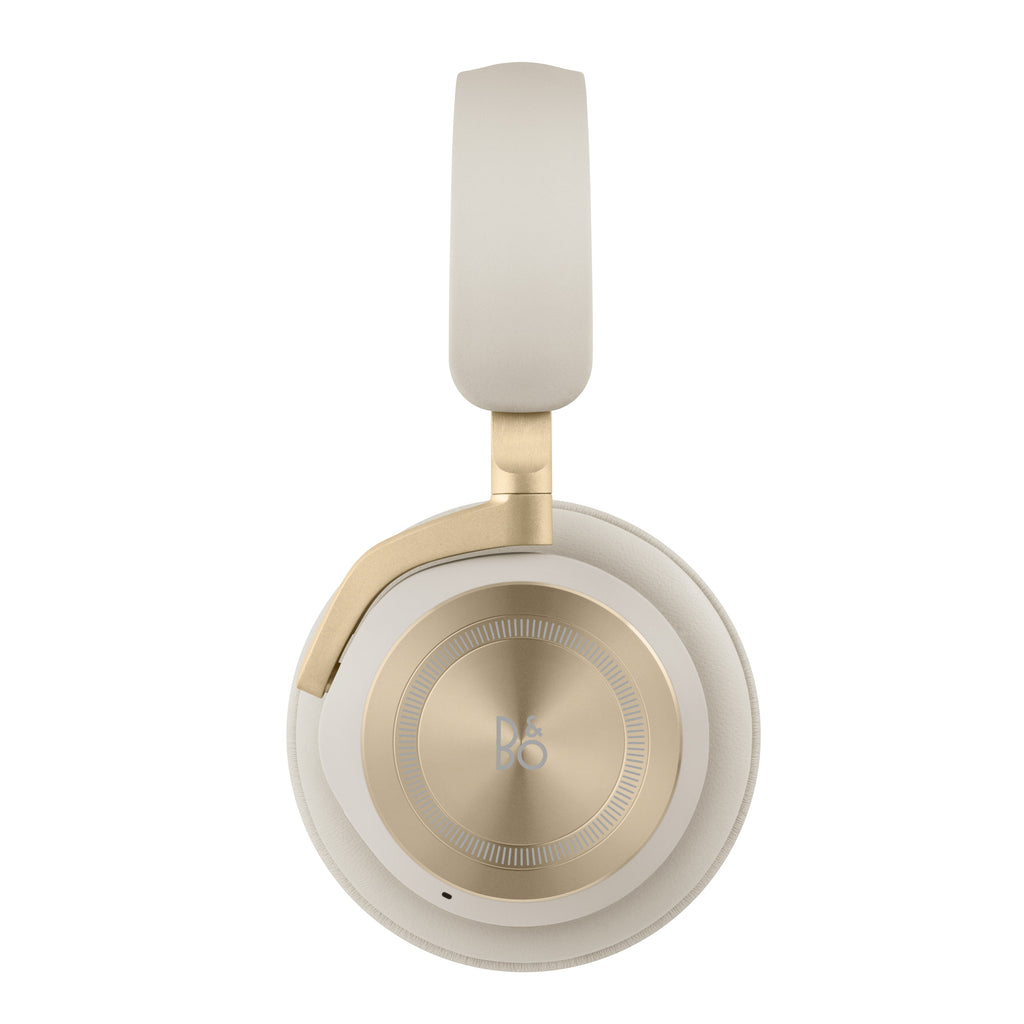 Seitenansicht des Bang & Olufsen Beoplay HX Kopfhörers in Sand: Fokus auf die kreisrunde, goldfarbene Aluminium-Ohrmuschel mit präziser Rillenstruktur, das dezente B&O-Logo und den passenden Lammleder-Kopfbügel.