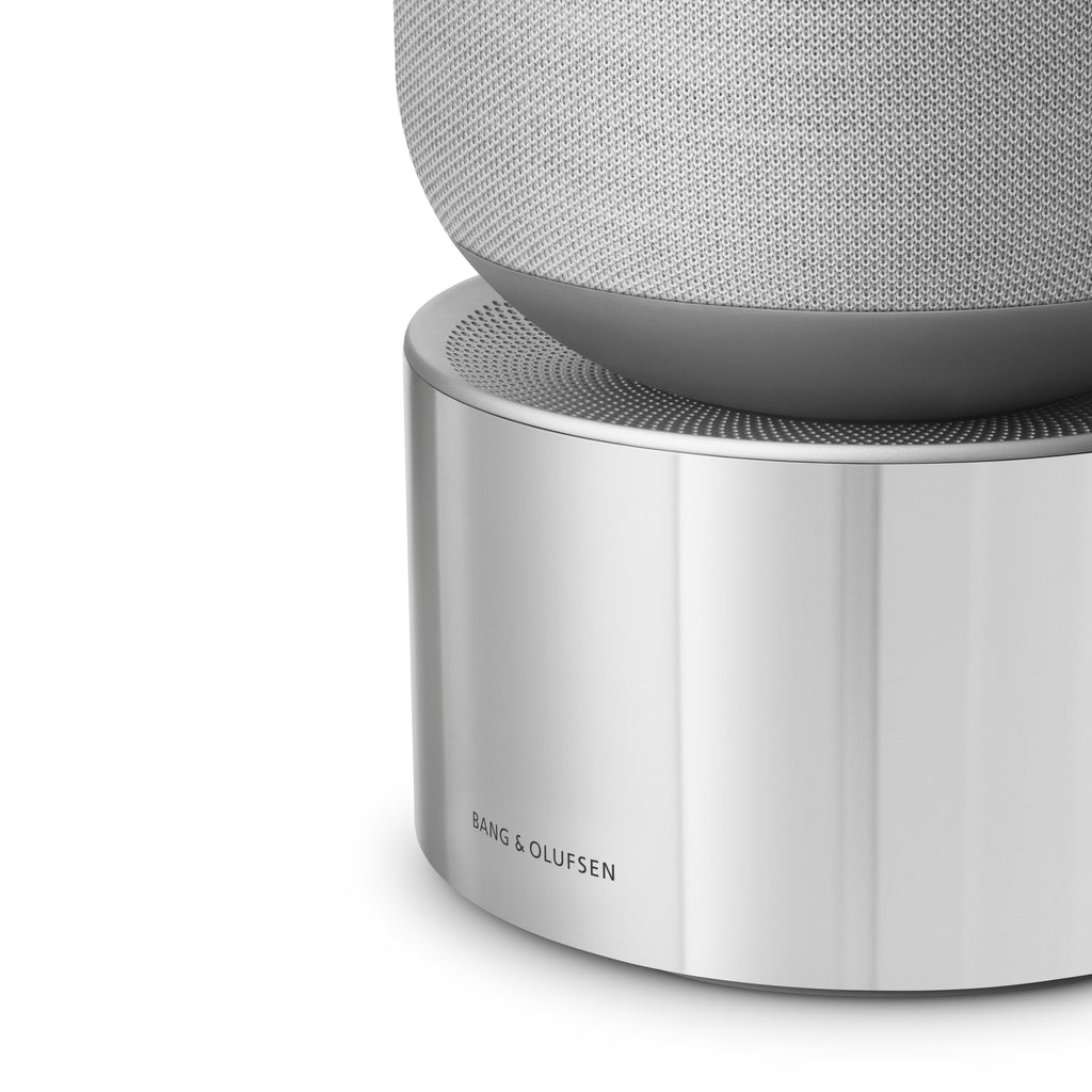 Detailaufnahme des Bang & Olufsen Beosound Balance in Natural Aluminium: Nahansicht der massiven, silbernen Aluminiumbasis mit dezenter Logo-Gravur und dem feinen Übergang zum grauen Kvadrat-Textilbezug sowie dem präzise gelochten Aluminium-Gitter.